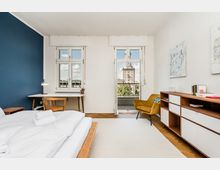 Helles, modern eingerichtetes Schlafzimmer mit bodennahem Bett, Schreibtisch am Fenster und Sideboard an der rechten Wand. Zwei hohe Fenster und eine Balkontür führen nach draußen; Parkettboden im Fischgrätmuster, blauer Akzentwandbereich und Blick auf ein markantes Gebäude in der Ferne.