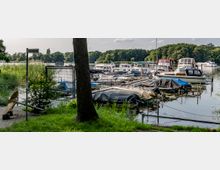 Hafenbereich an einer Seepromenade mit mehreren festgemachten Motor- und Segelbooten, teilweise abgedeckt, umgeben von Schilf und Bäumen im Hintergrund. Ein Gehweg mit Rasenfläche, ein großer Schiffspropeller und ein Schild mit der Aufschrift „Seepromenade“ sind im Vordergrund zu sehen.
