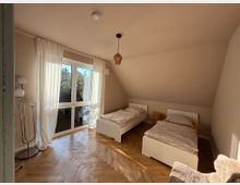 Schlafzimmer mit zwei einzelstehenden Betten und Holzfußboden in Fischgrätmuster. Ein großes Fenster mit lichtdurchlässigen Vorhängen bietet Blick auf eine grüne Außenumgebung, und eine Stehlampe steht in der Ecke.