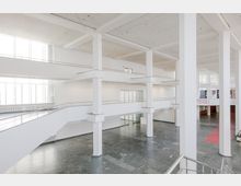 Heller, moderner Innenraum eines mehrstöckigen Gebäudes mit großen weißen Wänden, breiten Säulen und offenen Galerien auf mehreren Ebenen. Der Boden besteht aus dunklen Steinfliesen, und große Fenster lassen viel Tageslicht herein.