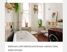 Badezimmer mit Badewanne, Toilette, Bidet und Waschmaschine, dekoriert mit bunten Fliesen an der Wand. Pflanzen auf Fensterbänken und Regalen bieten eine grüne Atmosphäre, ergänzt durch Holzelemente und natürliche Beleuchtung.