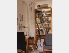 Ein gemütlicher Innenraum mit einem Holzregal voller Bücher, umgeben von einer leicht abgenutzten, verputzten Wand. Ein Schreibtisch mit Lampe und Stuhl befindet sich links vom Regal, und im Vordergrund steht ein gepolsterter Sessel.