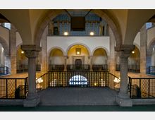 Innenansicht eines Gebäudes mit romanischen Elementen, darunter Rundbögen, verzierte Säulen und ein Metallgeländer im Vordergrund. Im Hintergrund befinden sich eine verzierte Tür, mehrere Arkaden und eine Balustrade, die auf eine mehrstöckige Struktur hindeuten.