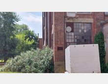 Alte Backsteinfassade eines Industriegebäudes mit großen, teilweise beschädigten Glasfenstern. Rechts ist ein zugewachsener Bereich mit Efeu und links Büsche und Bäume; im Vordergrund steht ein weißer Anhänger.