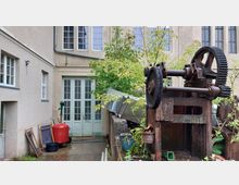 Hinterhof mit alten Maschinenkomponenten, darunter ein rostiges Zahnradgerät im Vordergrund. Im Hintergrund befindet sich eine beige Gebäude-Fassade mit großen Fensterflächen und einer Tür mit der Aufschrift „Privat“, umgeben von Pflanzen und verschiedenen Gegenständen.