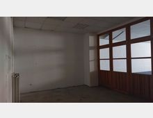 Leerer Raum mit weißen Wänden und grauem Boden. Auf der rechten Seite befinden sich Holztüren mit großen Glaseinsätzen, die in einen angrenzenden Bereich mit weiß gestrichener Ziegelwand blicken lassen.