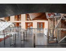 Modernes Gebäudeinnere mit Glas- und Metallarchitektur. Sichtbar sind hohe Glasfenster, Holzpaneele, eine gewölbte Holzdecke sowie skulpturale Metallstrukturen; im Vordergrund stehen mehrere runde Hochtische auf einem dunklen Boden.