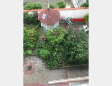 Blick auf einen begrünten Innenhof mit einem kleinen achteckigen Pavillon mit roten Dachziegeln und Solarzelle. Umgeben von Pflanzen, Blumenbeeten und rechts einem Abstellbereich mit mehreren Fahrrädern und weißen Gartenstühlen vor einer gemauerten weißen Wand.