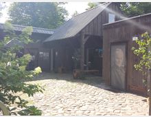 Holzhütten mit einer gepflasterten Hoffläche, umgeben von Bäumen. Eine Tür zeigt das Porträt einer Frau, daneben Pflanzen und Sitzmöglichkeiten; im Hintergrund sind Menschen zu sehen, die den Hof betreten.