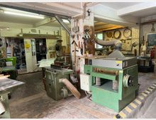 Werkstatt mit Holzmaschinen und Werkzeugen, darunter eine grüne Hobelmaschine im Vordergrund. Die Umgebung umfasst Werkbänke, Wände mit aufgehängten Werkzeugen und Schläuche zur Staubabsaugung, während im Hintergrund eine Tür zu einem weiteren Raum führt.