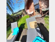 Ein Gartenbereich mit zwei grauen Liegestühlen auf einer Holzplattform, daneben eine holzverkleidete Sauna in Fassform mit geöffneter Tür. Im Hintergrund sind grüne Wiese, Bäume und ein Zaun zu sehen.