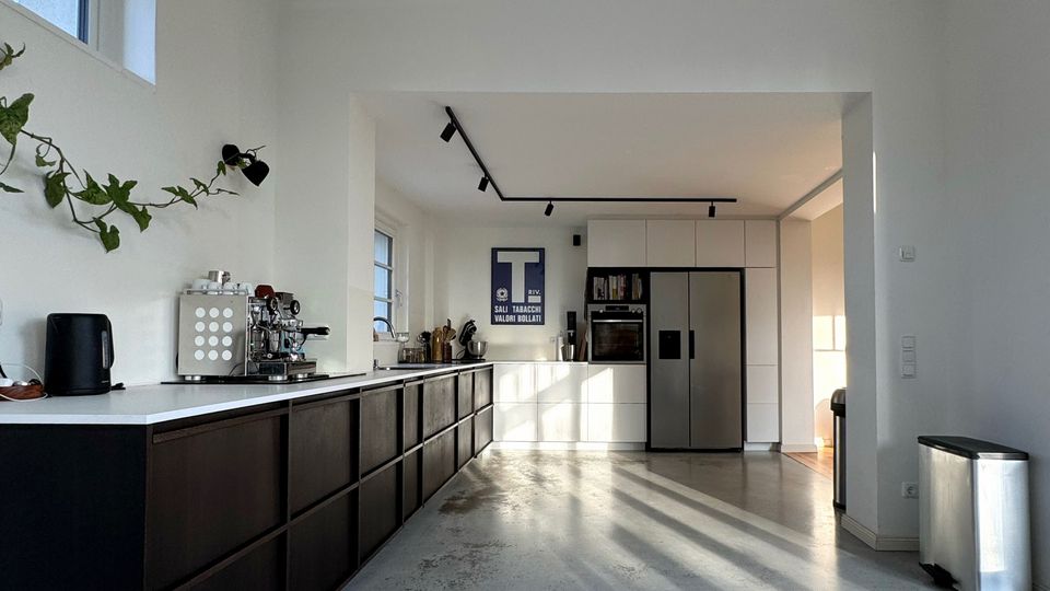 Moderne, offene Küche mit langen dunklen Unterschränken und heller Arbeitsplatte, Betonboden und weißen Wänden. Im Hintergrund stehen hohe Schränke mit Einbaugeräten und ein Edelstahl-Kühlschrank; Deckenstrahler und ein großes seitliches Fenster sorgen für Licht.