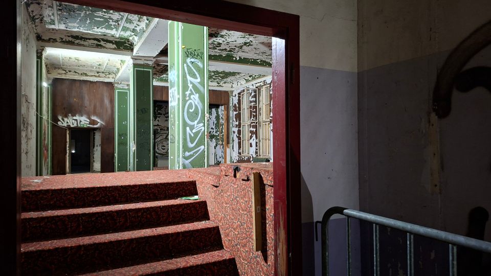 Blick durch einen Türrahmen in das Innere eines verlassenen Gebäudes mit rot gemusterten Stufen, abblätternder Deckenfarbe und grünen, teils mit Graffiti bedeckten Pfeilern. Im Hintergrund sind alte Holzverkleidungen, Fenster und stark verwitterte Wände zu sehen.