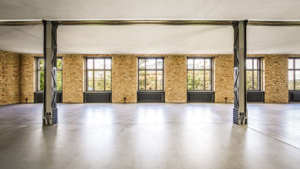 Großer, leerer Loft- oder Industriehallenraum mit glattem Betonboden, sichtbaren Stahlstützen und einer langen Backsteinwand. Mehrere hohe Sprossenfenster mit Heizkörpern darunter lassen viel Tageslicht in den Raum.