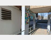 Überdachter Durchgang an einem Gebäude mit weiß verputzten Wänden und Stützen, versehen mit einem bunten Kachelmosaikband entlang der Decke. Ein gepflasterter Weg mit Metallgeländern führt durch den Gang; im Hintergrund sind ein Hofbereich, Bäume und weitere Wohngebäude zu sehen.