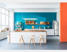 Moderne Küche mit einer weißen Kücheninsel mit Holzplatte, drei Holz-Barhockern und einer blauen Wand im Hintergrund. Offene Regale, ein Hängeregal, ein Kühlschrank aus Edelstahl sowie ein großes Fenster auf der linken Seite prägen den Raum.