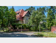 Eckhaus im Stil einer Altbauvilla mit hellem Putz, rotem Ziegeldach und Fachwerkdetails, umgeben von Bäumen und einer niedrigen roten Ziegelmauer; im Vordergrund eine gepflasterte Straßenecke mit Straßenschildern.