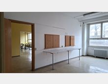 Leerer Innenraum in einem älteren Gebäude mit weißen Wänden, einer langen schmalen Ablage auf Metallbeinen und zwei an der Wand montierten Holzschränken. Rechts befindet sich ein großes Fenster mit Heizkörper darunter, links ein offener Türrahmen mit Blick in einen Nebenraum mit Tischen.