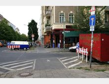 Straßenecke in einem Wohngebiet mit einem mehrstöckigen Altbau und einem Café im Erdgeschoss auf der rechten Seite. Darauf sind Straßenschilder, Absperrungen, ein geparkter Oldtimer-Van, sowie Bäume und Menschen auf dem Gehweg sichtbar.