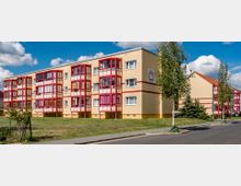 Mehrfamilienhaus mit gelber Fassade und roten Balkonen, umgeben von kleinen Grünflächen und einer asphaltierten Straße im Vordergrund, sowie vereinzelt stehenden Bäumen. Im Hintergrund ist ein wolkenloser Himmel zu sehen.