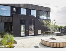 Modernes Gebäude mit dunkler Holzfassade und großen Fenstern. Im Vordergrund ein gepflasterter Innenhof mit Sitzbänken aus Holz, einem Baum in einer runden Holzumrandung und bepflanzten Beeten.