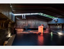 Ein rustikaler Innenraum mit hölzernen und metallischen Oberflächen, genutzt für eine Theater- oder Performance-Aufführung. Eine Person in einem weißen Kleid steht auf einer dunklen Bühne, während zwei weitere Personen in weißen Kostümen an den oberen Strukturen hängen oder lehnen, beleuchtet von farbigen Lichtern.