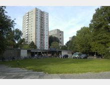 Wohnblock-Siedlung mit einem mehrstöckigen Hochhaus im Hintergrund, umgeben von Bäumen und Grünflächen. Im Vordergrund befinden sich ein Eingang zur U-Bahn-Station mit Fahrradständern sowie parkende Fahrzeuge, darunter ein Polizeifahrzeug.