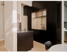 Moderne, minimalistische Küche mit mattschwarzen Einbauschränken und einer großen Nische aus Edelstahl mit Spüle und Armatur. Helle Wände und ein Holzfußboden rahmen die Küchenzeile ein, daneben steht eine kleine Insel mit Edelstahl-Arbeitsplatte.