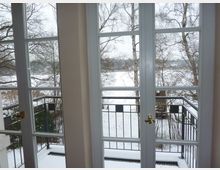 Blick durch bodentiefe Fenster auf einen kleinen Balkon mit dunklem Metallgeländer, dahinter eine verschneite winterliche Landschaft mit kahlen Bäumen und Wiesen.