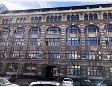 Fassade eines großen, mehrstöckigen historischen Gebäudes mit grauem Stein, hohen Rundbogenfenstern und reich verzierten Details. Im Erdgeschoss befindet sich ein Durchgang, daneben sind parkende Autos zu sehen.