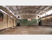 Verlassene Turnhalle mit hohem, gewölbtem Betondeckengebälk und großen Fensterbändern entlang der Seitenwände. An der Stirnwand hängt ein Basketballkorb vor abblätternder grüner Farbe, der Boden ist staubig und beschädigt, mit offenen Türdurchgängen an beiden Seiten.