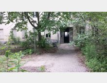 Ein verwilderter Eingangsbereich eines verlassenen Gebäudes mit offenen Türen, umgeben von dichtem Bewuchs und Bäumen. Der gepflasterte Weg ist von Vegetation überwuchert, die Fassade zeigt geschlossene Fensterläden.