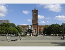 Rathausplatz mit dem markanten roten Backsteingebäude des Berliner Rathauses im Hintergrund, davor der Neptunbrunnen mit Figuren und Wasserfontänen, umgeben von Bäumen und Passanten auf dem gepflasterten Platz.
