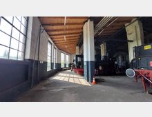 Ein Innenraum eines historischen Lokschuppens mit hohen Fenstern auf der linken Seite, die viel Tageslicht hereinlassen. Holzbalkendecke, weiße Wände mit dunkelgrauer Unterseite und mehrere alte Dampfloks und Fahrzeuge sind zu sehen.