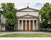 Neoklassizistische Kirchenfassade mit einem Säulenportal und dreieckigem Giebel, umgeben von hohen Bäumen. Die Fassade ist aus hellem Stein, die Türen bestehen aus Holz, und der Vorplatz ist mit Rasen und Pflaster gestaltet.