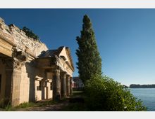 Ruine eines historischen Gebäudes mit verzierten Säulen und teilweise eingestürztem Dach, umgeben von Büschen und Bäumen. Im Hintergrund ist ein Fluss mit bewaldetem Ufer unter klarem, blauem Himmel zu sehen.