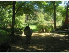Gartenbereich mit Steinboden und Gartenmöbeln, umgeben von Bäumen und Pflanzen. Im Hintergrund Wiese mit Zaun, Blumentöpfen und einem dekorativen Holzgestell.