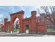 Backsteinbogen und -türme am Eingang der Technischen Hochschule Brandenburg. Der Eingang wird von roten Ziegelmauern eingerahmt, mit einem grünen Metalltor und Bäumen im Hintergrund.