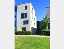 Moderne, mehrstöckige Wohngebäude mit heller Fassade und großen Fenstern, umgeben von Grünflächen, Hecken und einem Gehweg; im Hintergrund blauer Himmel.