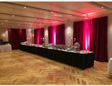 Ein großer Saal mit hölzernem Parkettboden, roten Vorhängen entlang der Wände und stimmungsvoller Beleuchtung. In der Mitte steht ein langes Buffet mit Geschirr, Speisen und silbernem Serviergeschirr, flankiert von Kerzenleuchtern.