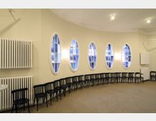 Ein heller Raum mit beigefarbenen Wänden und ovalen Fenstern, die in einer Reihe entlang einer leicht gebogenen Wand angeordnet sind. Entlang der Wand stehen schwarze Holzstühle, und auf der linken Seite sind weiße Heizkörper montiert.