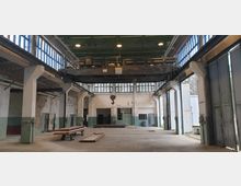 Innenansicht einer leerstehenden Industriehalle mit hohen Decken, großflächigen Fenstern und Stahlträgern. In der Mitte hängt ein Kranausleger, während an den Seiten Türen und Fenster mit teilweise verwitterten Wänden sichtbar sind.