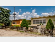 Flaches Gebäude mit wellenförmigem Dach und gelb-orange gestrichenen Wänden, umgeben von gepflegter Bepflanzung und einer Holzzaunbegrenzung. Im Hintergrund ein hoher Sendeturm vor einem blauen Himmel mit weißen Wolken.