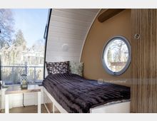 Ein kleines, modernes Holzpod mit abgerundeter Decke, einem seitlichen Bullaugenfenster und einer großen Glasfront, die auf ein bewachsenes Außenumfeld blickt. Innen ein Bett mit dunkler Decke, gemusterten Kissen und einem kleinen Tisch mit Laterne.