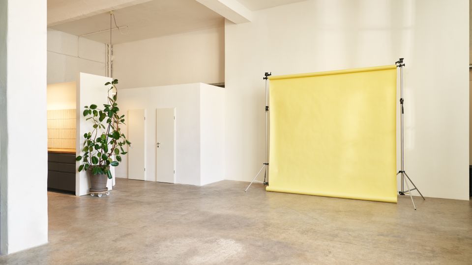 Heller, minimalistischer Studio- oder Arbeitsraum mit Betonboden und weißen Wänden, darunter ein abgetrennter Kubus mit zwei weißen Türen. Rechts steht ein gelber Fotohintergrund auf Stativgestellen, links eine große Topfpflanze neben einem niedrigen Schrank.