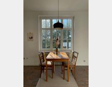 Ein heller Essbereich mit einem rechteckigen Holztisch und vier Stühlen, vor einer großen Sprossenfensterfront. Eine schwarze Hängeleuchte hängt über dem Tisch, der mit einer Vase und kleinen Gegenständen dekoriert ist.