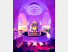 Innenraum einer modernen umgebauten Kirche mit gotischen Spitzbögen, beleuchtet in violettem Licht. Das Zentrum bildet ein Altarbereich mit farbigem Glasfenster, umgeben von Sitzgelegenheiten, Kissen und geschwungenen Plattformen.