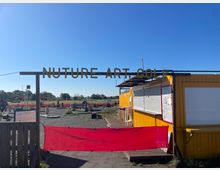 Ein Minigolfplatz unter freiem Himmel mit künstlerischen Elementen, erkennbar an der Beschriftung "Nuture Art Golf". Im Vordergrund ein gelber Container mit geschlossenen Rollläden, daneben eine rote Absperrung; im Hintergrund Spielflächen und offene Landschaft.