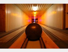 Ansicht einer Kegelbahn aus niedriger Perspektive, mit einer dunklen Kegelkugel im Vordergrund auf einer Schiene. Der lange Gang ist von warmem, gelbem Licht beleuchtet, mit seitlichen Holzverkleidungen und bunten Lichteffekten am Ende der Bahn.