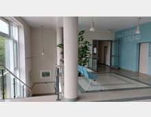 Heller Flur in einem modernen Gebäude mit beigen und blauen Wänden, großen Fenstern und einer Treppe mit einem Glasgeländer. Es gibt eine blaue Sitzbank, Zimmernummern an der Wand und grüne Pflanzen zur Dekoration.